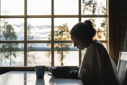 Finnland, Lappland, junge Frau sitzt am Fenster an einem See und schaut in ihr Tagebuch - KKAF02343