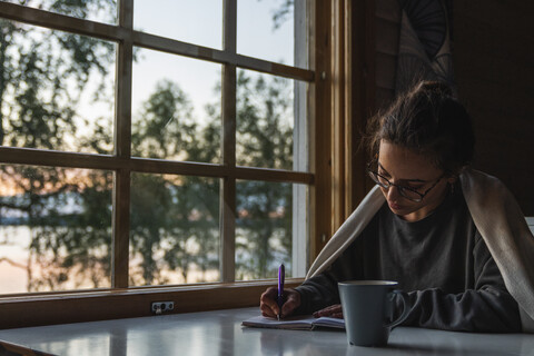 Finnland, Lappland, junge Frau sitzt am Fenster an einem See und schreibt in ihr Tagebuch, lizenzfreies Stockfoto