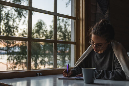 Finland, Lapland, young woman sitting at the window at a lake writing into diary - KKAF02344