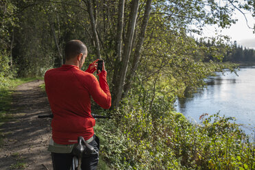 Mountainbiker steht am See und fotografiert mit seinem Smartphone - KKAF02359