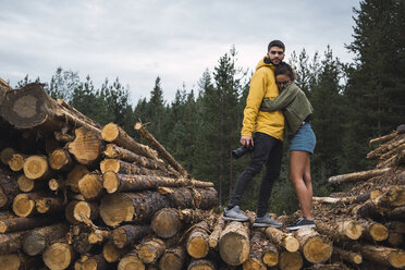 Junge Frau umarmt Mann mit Kamera auf Holzstapel - KKAF02385