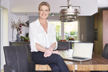 Porträt einer lächelnden Geschäftsfrau, die zu Hause auf einem Tisch mit einem Laptop sitzt - PDF01761