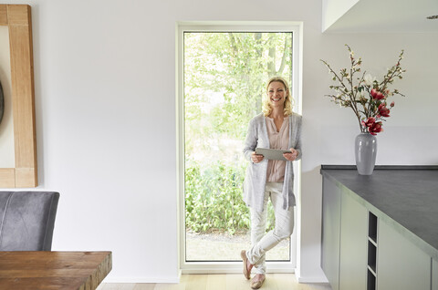 Porträt einer glücklichen Frau, die zu Hause an der französischen Tür steht und ein Tablet benutzt, lizenzfreies Stockfoto