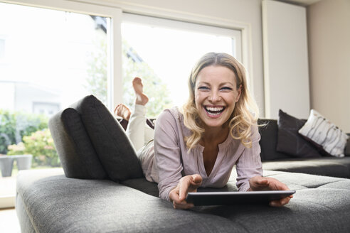 Porträt einer lachenden Frau, die zu Hause auf der Couch liegt und ein Tablet benutzt - PDF01780