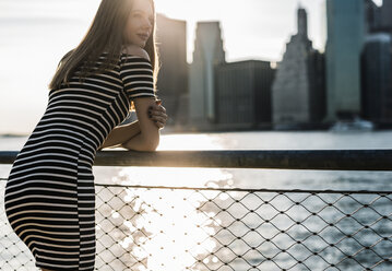 USA, New York, Brooklyn, woman wearing striped dress standing in front of East River by sunset - GIOF04555