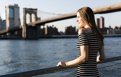 USA, New York, Brooklyn, woman wearing striped dress standing in front of East River by sunset - GIOF04557