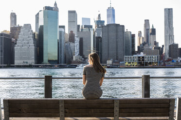 USA, New York, Brooklyn, Rückenansicht einer Frau, die auf einer Bank vor dem East River und der Skyline von Manhattan sitzt - GIOF04569