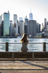 USA, New York, Brooklyn, Rückenansicht einer Frau, die auf einer Bank vor dem East River und der Skyline von Manhattan sitzt - GIOF04570