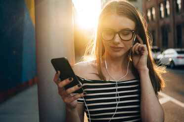 USA, New York, Brooklyn, Dumbo, portrait of woman listening music with smartphone and earphones at sunset - GIOF04597