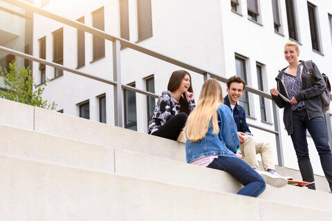 Vier junge erwachsene Studenten sitzen plaudernd auf einer Treppe vor dem College, Blick aus geringer Höhe, lizenzfreies Stockfoto