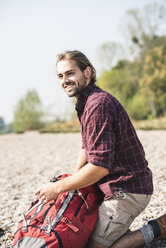 Lächelnder junger Mann mit Rucksack am Kieselsteinufer - UUF15312