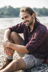 Smiling young man sitting at the riverside - UUF15321