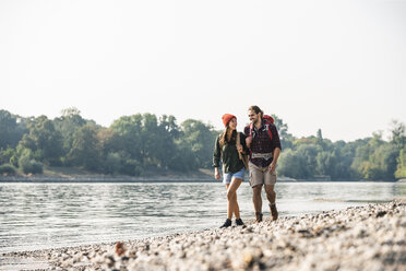 Lächelndes junges Paar mit Rucksäcken beim Spaziergang am Flussufer - UUF15325