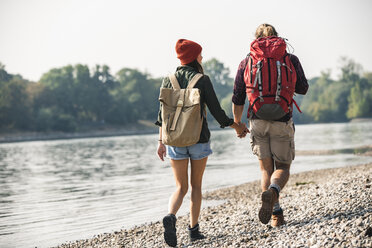 Rückansicht eines jungen Paares mit Rucksäcken, das Hand in Hand am Flussufer spazieren geht - UUF15326