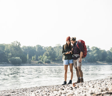 Junges Paar mit Rucksäcken am Flussufer, das sein Mobiltelefon überprüft - UUF15328