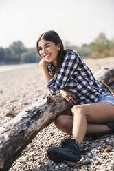 Glückliche junge Frau sitzt an einem Baumstamm am Kiesstrand - UUF15342