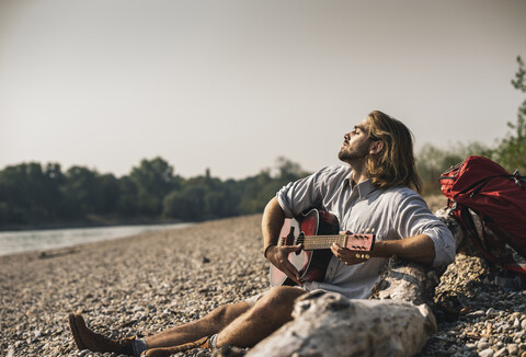 Junger Mann sitzt am Flussufer und spielt Gitarre, lizenzfreies Stockfoto