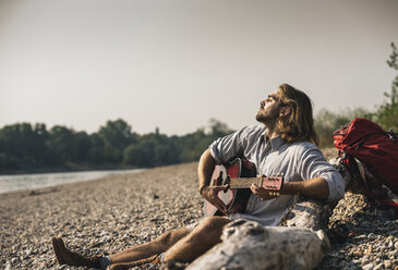 Young man sitting at the riverside playing guitar - UUF15350