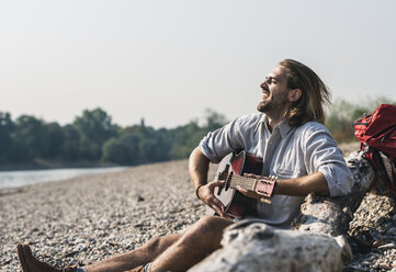 Übermütiger junger Mann sitzt am Flussufer und spielt Gitarre - UUF15351