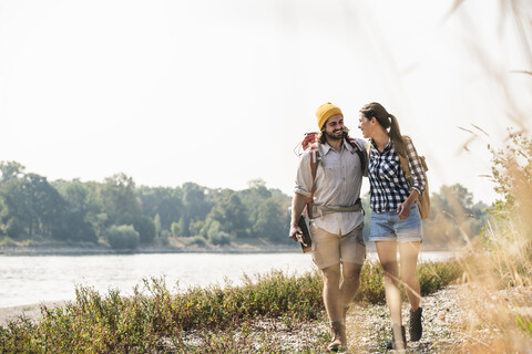Glückliches junges Paar mit Rucksäcken und Gitarre beim Spaziergang am Flussufer, lizenzfreies Stockfoto