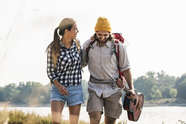 Happy young couple with backpacks and guitar walking at the riverside - UUF15359