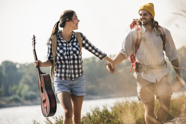 Happy young couple with backpacks and guitar walking at the riverside - UUF15360