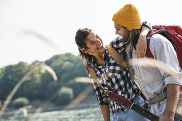 Happy young couple with backpacks and guitar embracing at the riverside - UUF15364