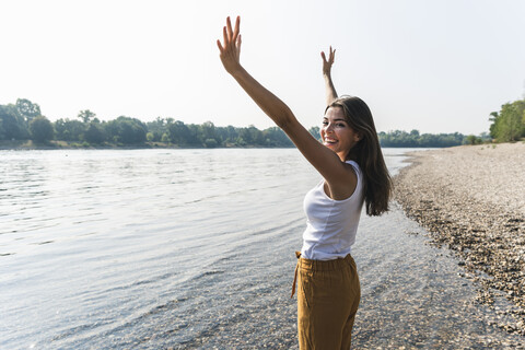 Glückliche junge Frau steht am Flussufer und hebt die Arme, lizenzfreies Stockfoto