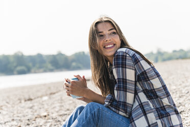 Happy young woman holding a mug sitting at the riverside - UUF15408