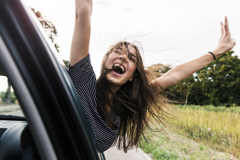 Unbekümmerte junge Frau lehnt sich schreiend aus dem Autofenster, lizenzfreies Stockfoto
