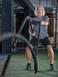 Athletischer reifer Mann beim Training mit Kampfseilen im Fitnessstudio - STSF01753