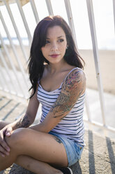 Porträt einer jungen Frau mit Nasenpiercing und Tattoos, die auf einem Balkon im Gegenlicht sitzt - GIOF04603