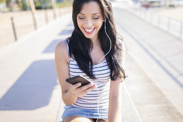 Happy young woman with nose piercing and tattoo using earphones and smartphone on promenade - GIOF04605