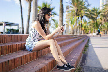 Spain, Barcelona, smiling young woman sitting on steps at sunlight using cell phone - GIOF04609