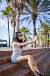 Spain, Barcelona, young woman sitting on steps at sunlight taking selfie with smartphone - GIOF04610