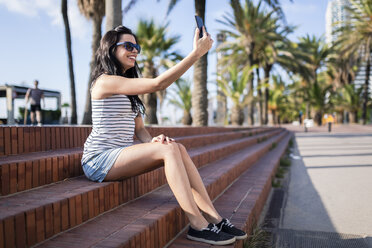 Spanien, Barcelona, junge Frau sitzt auf Stufen im Sonnenlicht und macht ein Selfie mit Smartphone - GIOF04611