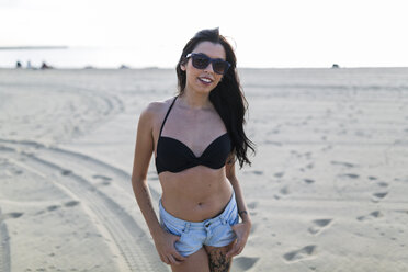 Portrait of smiling young woman wearing bikini top and hot pants standing on the beach - GIOF04620
