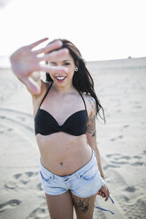 Porträt einer lächelnden jungen Frau mit Nasenpiercing und Tattoo, die am Strand durch ihre Finger schaut - GIOF04623