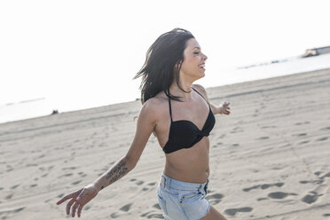 Glückliche junge Frau mit Tattoo und Nasenpiercing läuft am Strand - GIOF04626