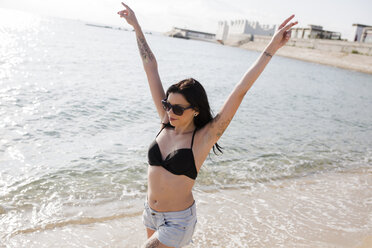 Happy young woman walking at seashore with arms raised - GIOF04629