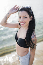 Porträt einer lächelnden jungen Frau mit Nasenpiercing und Tattoos, die am Meer steht - GIOF04632
