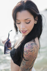 Portrait of smiling young woman with nose piercing and tattoo on her shoulder at the sea - GIOF04633