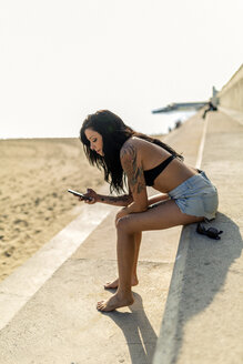 Lächelnde junge Frau mit Nasenpiercing und Tattoos, die ihr Smartphone in der Nähe des Strandes benutzt - GIOF04636