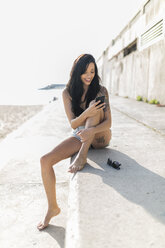 Lächelnde junge Frau mit Nasenpiercing und Tattoo, die ihr Smartphone in der Nähe des Strandes benutzt - GIOF04637