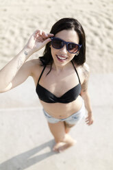 Porträt einer lächelnden jungen Frau mit Nasenpiercing und Tattoos, die am Strand eine Sonnenbrille trägt - GIOF04639