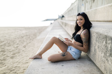Portrait of tattooed young woman reading a book near the beach - GIOF04643