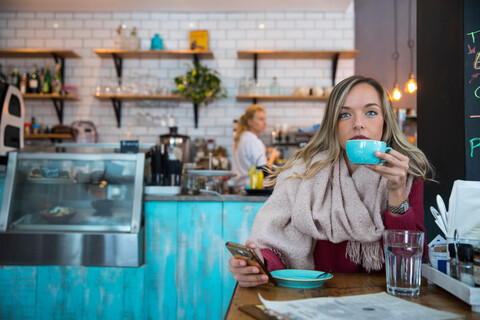 Frau sitzt in einem Café, hält ihr Smartphone und trinkt Kaffee, lizenzfreies Stockfoto