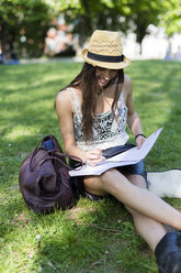 Lächelnde junge Studentin, die auf einer Wiese in einem Park sitzt und ein Schreibheft benutzt - GIOF04682