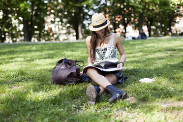 Lächelnde junge Studentin, die auf einer Wiese in einem Park sitzt und ein Schreibheft benutzt - GIOF04687