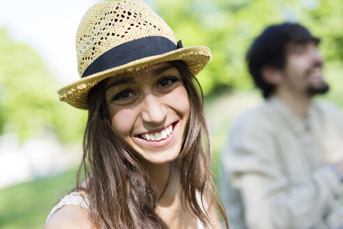 Porträt einer glücklichen jungen Frau in einem Park mit ihrem Freund im Hintergrund - GIOF04692